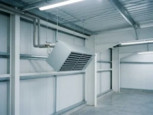Lichtgrijze unit heater opgehangen aan stalen gordingen onder een golfplaten dak, met neerwaartse luikerrooster en pijpaansluitingen bovenop.