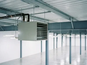 Lichtgrijze luchtverwarmer hangend aan staalconstructie, met geribbeld metalen plafond, neerwaartse uitblaasrooster en pijpaansluitingen bovenop.