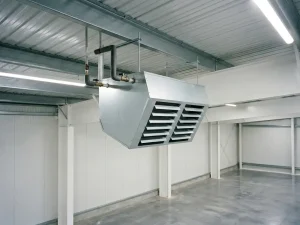 Lichtgrijze luchtverwarmer opgehangen aan stalen gordingen bij een golfplafond, met neerwaartse lamellengrill en leidingaansluitingen bovenop, in een industriële ruimte.
