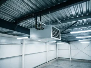 Lichtgrijze unit heater opgehangen aan stalen gordingen onder golfstalen dak, met neerwaartse lamellengrill en bovenste pijpaansluitingen.