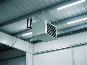 Lichtgrijze unit heater hangend aan stalen spanten nabij golfplaten plafond, met neerwaartse lamellengrill en bovenste pijpaansluitingen.