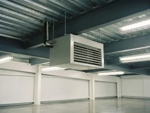 Lichtgrijze luchtverwarmer hangend aan stalen gordingen nabij golfplaten plafond, met geribbeld uitlaatrooster en pijpaansluitingen bovenop.